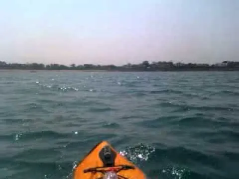 The bow of an orange kayak on choppy sea, with the low Guernsey coastline on the horizon.
